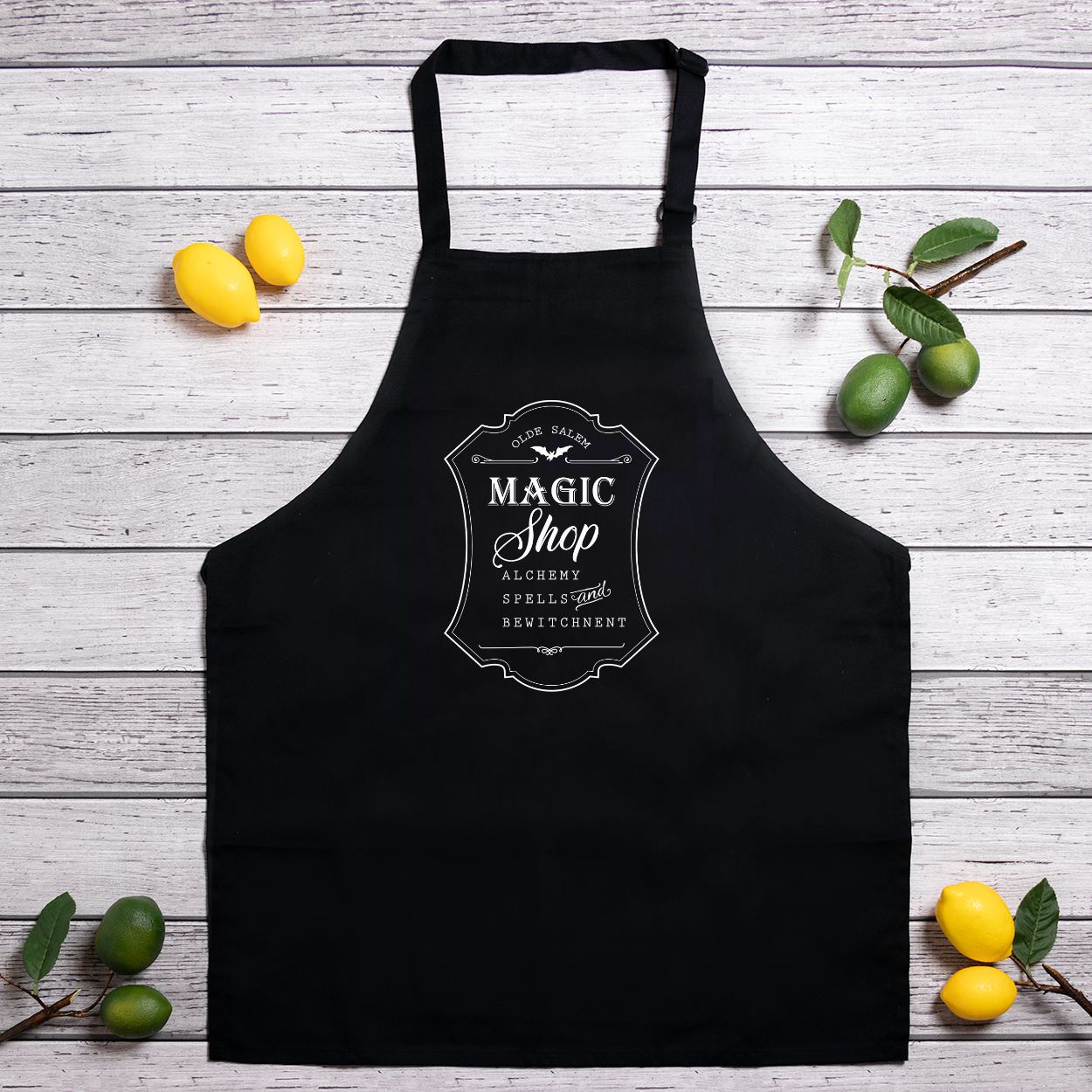Black apron with 'Magic Shop' design on a wooden surface with lemons and leaves.